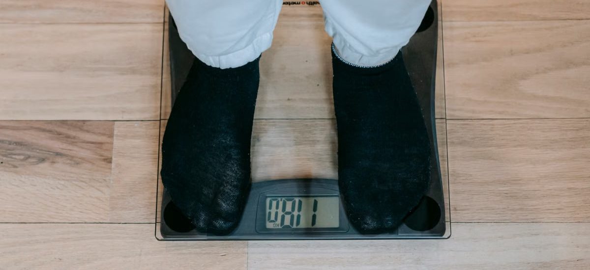 From above of crop unrecognizable plus size person in casual clothes standing on weighing scale on wooden floor