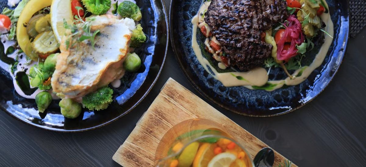 Top view of gourmet fish and steak dishes with fresh citrus and herbs on a stylish table setting.